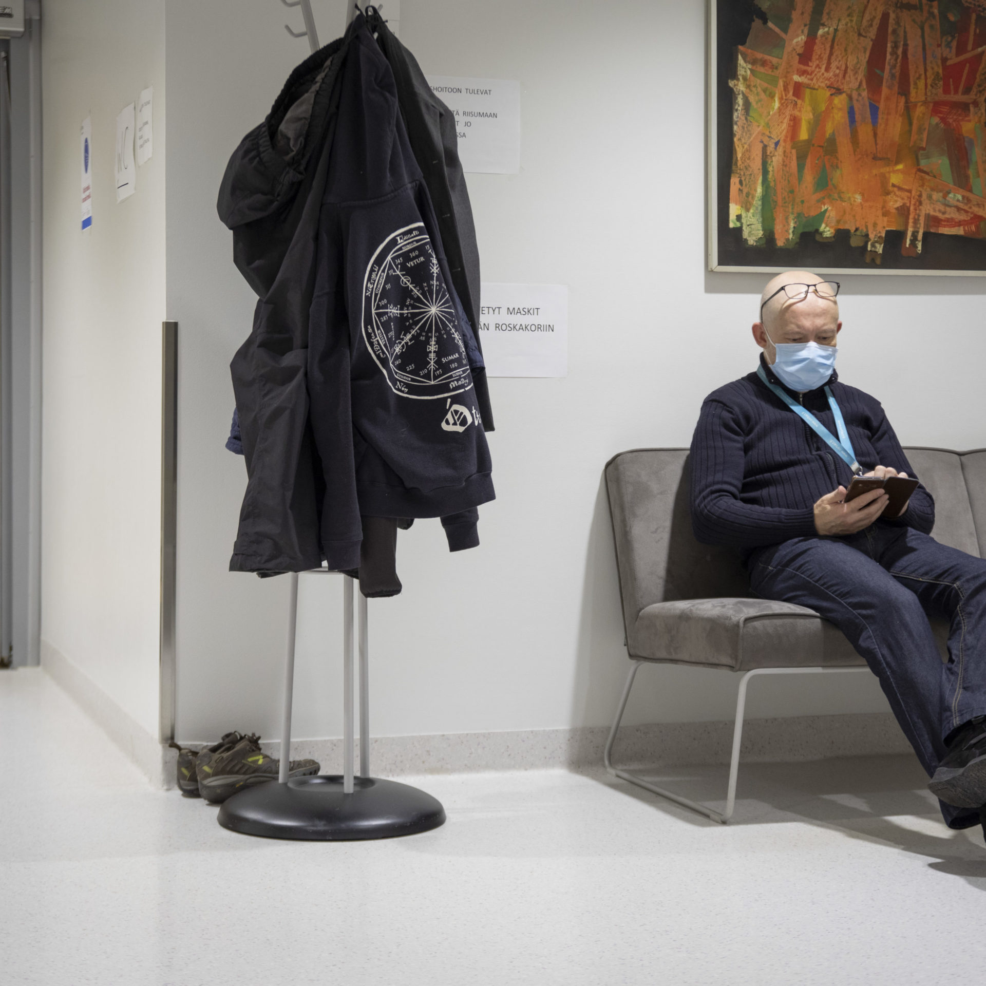Eläkeläinen istuu ja selaa puhelinta terveyskeskuksen odotustilassa.
