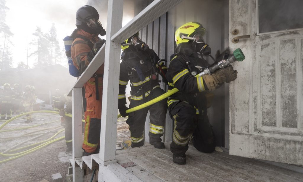 Kolme pelastajaa talonpolttoharjoituksessa savuavan talon ovella vesiletkun kanssa