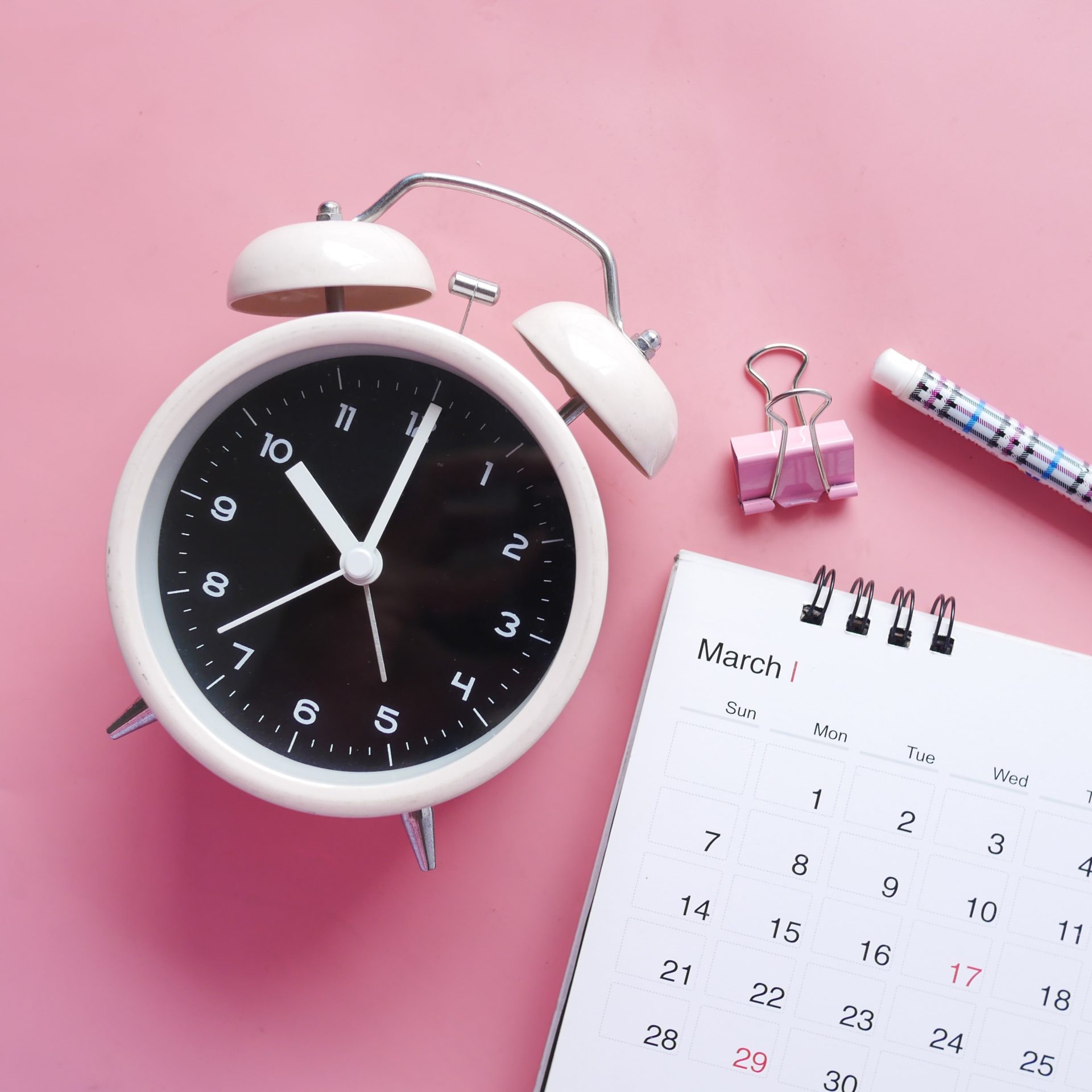 Herätyskello, kalenteri ja kynä vaaleanpunaisella taustalla